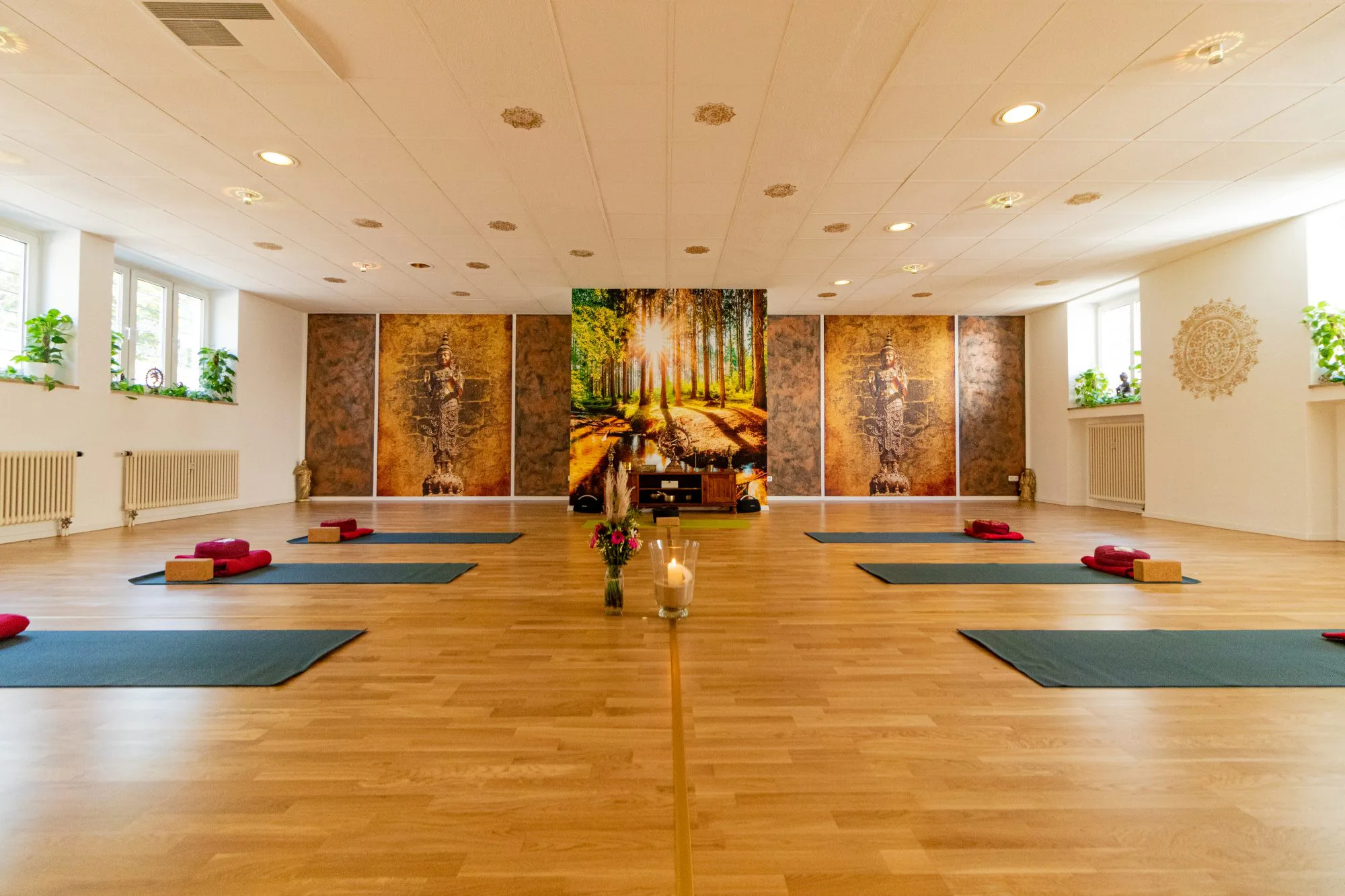 Yoga-Center-Würzburg-Glücksbringer Ein helles Yogastudio mit Holzböden, ordentlich aufgereihten Matten und Kissen heißt Sie willkommen. In der Mitte stehen Kerzen und Blumen, und an der Rückwand ziert ein farbenfrohes Waldwandbild den Raum. Als Teil unseres Zentrums für Wellness eignet sich dieser einladende Raum ideal für Gruppenkurse oder Teilnehmer der Yogalehrer-Ausbildung. Die Wände sind mit Fenstern gesäumt, durch die reichlich Tageslicht hereinströmt.