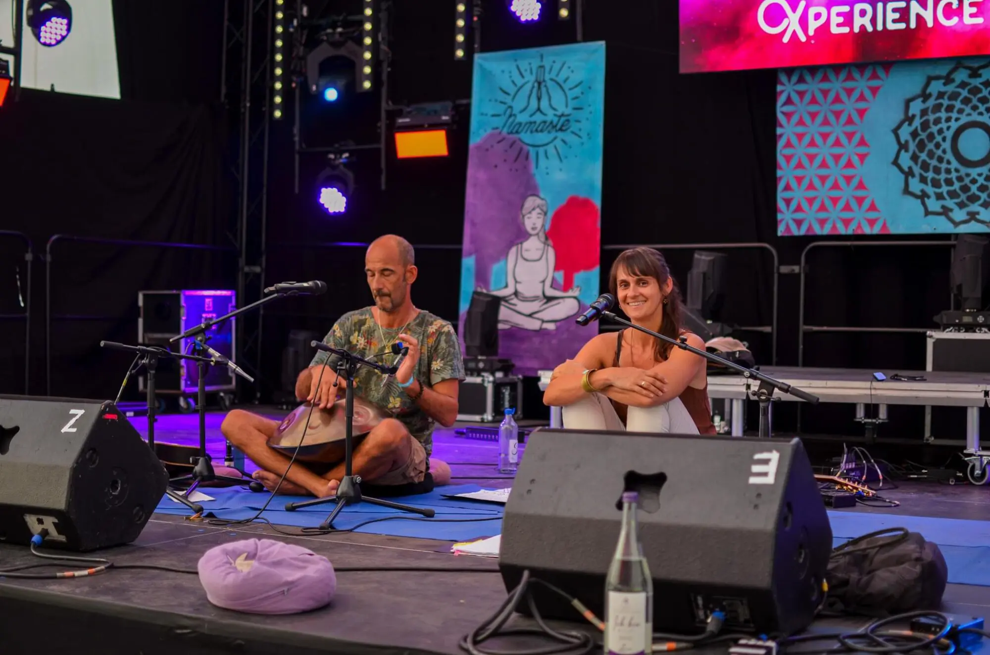 Bei einer Veranstaltung in der Yoga-Akademie in Würzburg sitzen zwei Personen mit Mikrofonen im Schneidersitz auf der Bühne, wobei eine von ihnen eine Handpan spielt. Im Hintergrund sind bunte Plakate, Bühnenbeleuchtung und Lautsprecher zu sehen, die eine lebendige Atmosphäre schaffen. Eine Wasserflasche und ein Kissen liegen auf dem Boden in ihrer Nähe und tragen zur entspannten Atmosphäre bei – perfekt für Momente der Besinnung, wie sie in Coaching-Ausbildungssitzungen oft vorkommen.