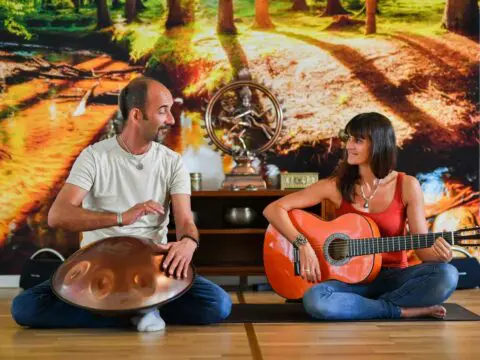 Ein Mann spielt auf einer Handpan, während eine Frau mit einer Gitarre neben ihm sitzt. Beide sitzen im Schneidersitz auf dem Boden eines gemütlichen Yogazentrums in Würzburg und lächeln sich vor dem Hintergrund eines farbenfrohen Waldwandbildes an.