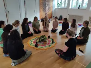 Gruppe von Menschen sitzt im Kreis auf dem Boden um ein dekoriertes Tuch mit Pflanzen und Figuren.