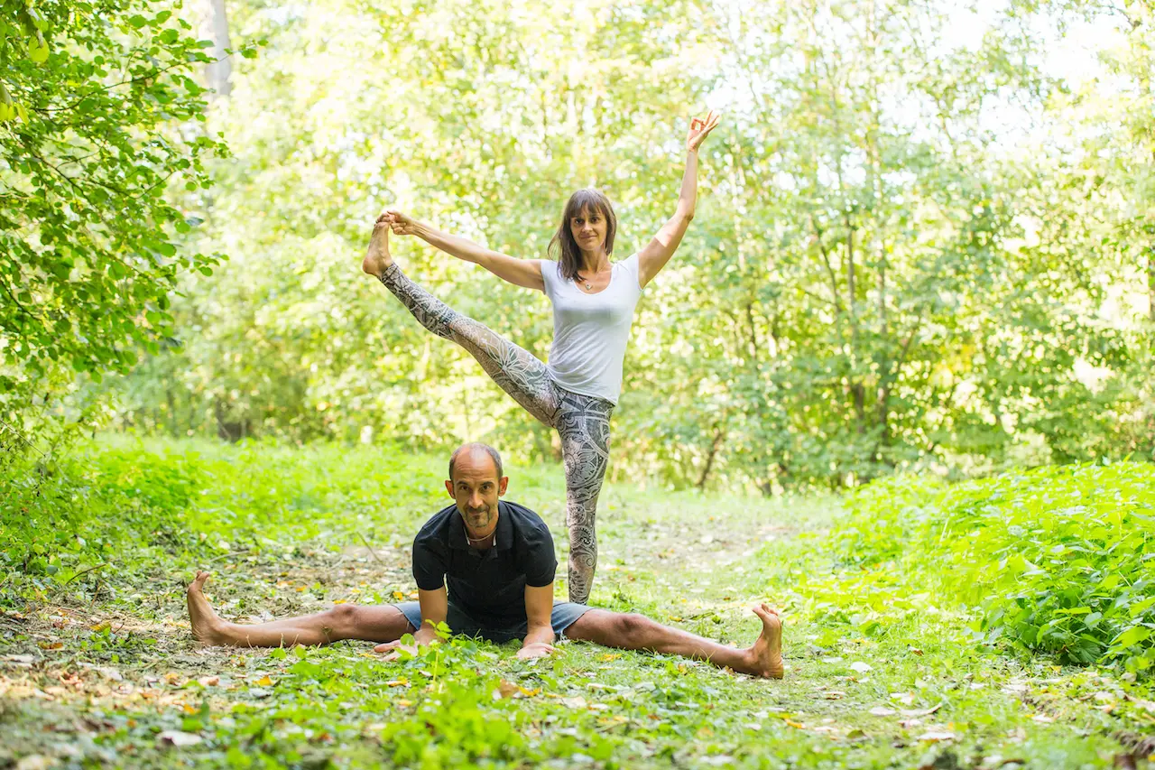 Ein Mann macht auf einem Waldweg einen Spagat, während eine Frau auf seinem Rücken balanciert, ein Bein hoch in die Luft streckt und mit einem erhobenen Arm lächelt. Beide tragen Sportkleidung in einer üppig grünen Naturkulisse – eine inspirierende Szene, die an fortgeschrittene Partnerübungen erinnert, wie man sie oft in einem Yogacenter oder bei Retreats der Yoga-Akademie sieht.